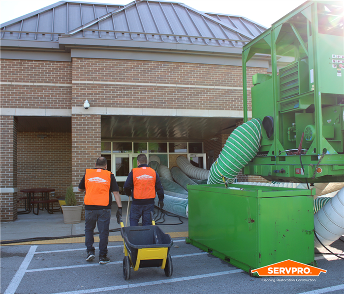 Two SERVPRO of Tampa Southeast employees in orange vests walking into a school next to a big, green desiccant dehumidifier 