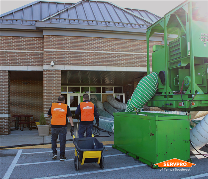 Two SERVPRO of Tampa Southeast crew in orange vests walking into a school next to a big, green desiccant dehumidifier 